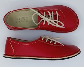 May include: A pair of red leather lace-up shoes with white laces. The shoes have a white rubber sole and the text "RUBY BAREFOOT" is printed on the inside of the shoe.