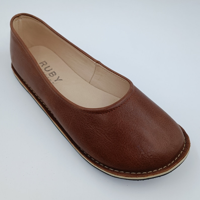 Peut inclure: Ballerine en cuir marron avec un bout arrondi. La chaussure a une semelle cousue et le mot "RUBY" est visible sur la semelle int&eacute;rieure. La chaussure est de couleur marron chaud.