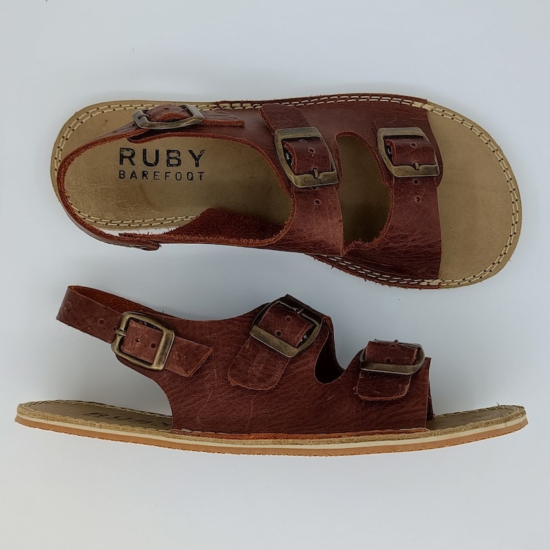 May include: A pair of brown leather sandals with two adjustable buckles on each strap. The sandals have a  "RUBY BAREFOOT" label on the top strap.