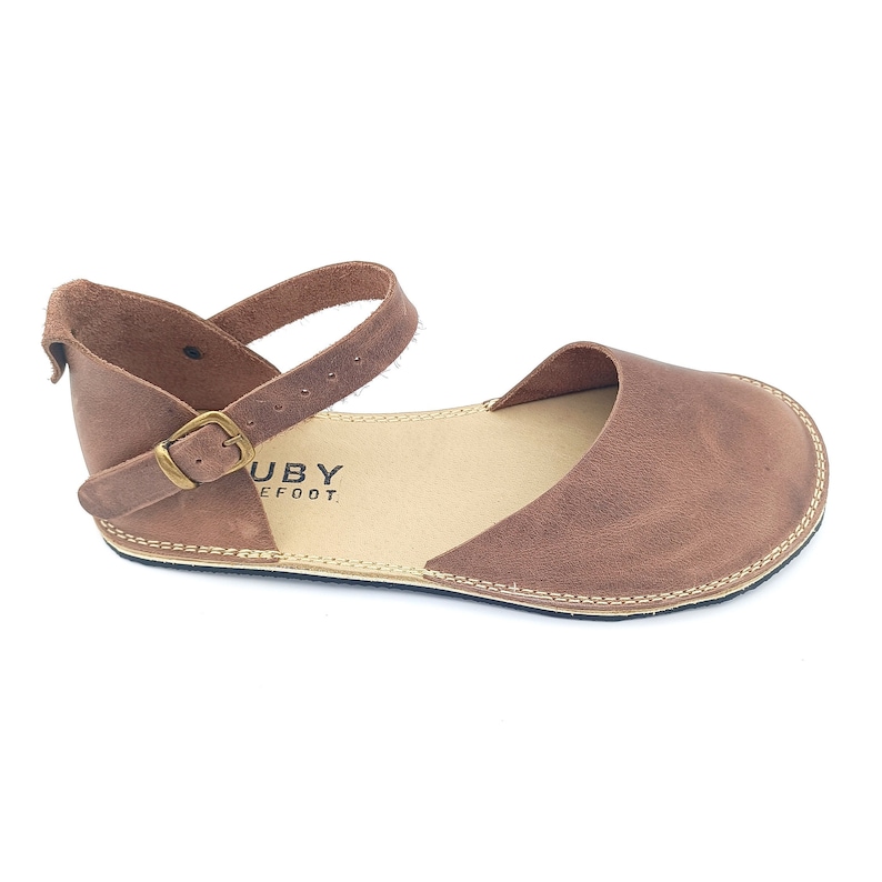 May include: Brown leather ankle strap sandals with a buckle closure. The sandals have a flat sole and are made by the brand "Ruby Barefoot."