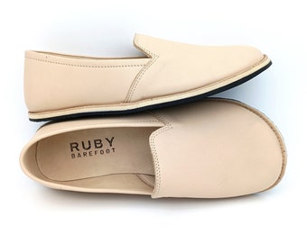 May include: A pair of light beige leather slip-on shoes with a black sole. The shoes have the text "RUBY BAREFOOT" printed on the insole.