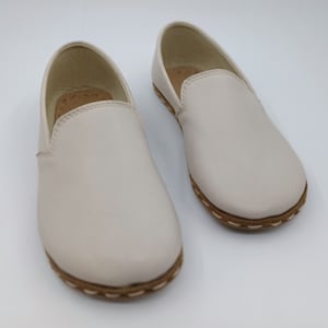 May include: A pair of off-white leather slip-on shoes with a brown stitched trim around the sole.