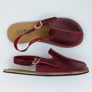 Może przedstawiać: Para burgundowych skórzanych sandałów z regulowanym paskiem z klamrą. Sandały mają logo "RUBY BAREFOOT" na wkładce.