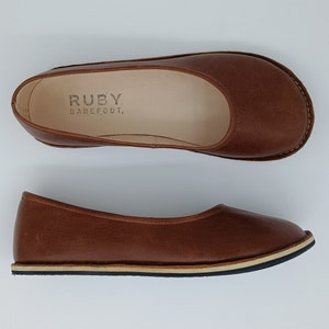 Peut inclure: Deux ballerines en cuir marron. Les chaussures ont un bout arrondi et une semelle cousue. La marque "RUBY BAREFOOT" est imprim&eacute;e sur la semelle int&eacute;rieure d'une chaussure. Les chaussures sont con&ccedil;ues pour le confort et le style.
