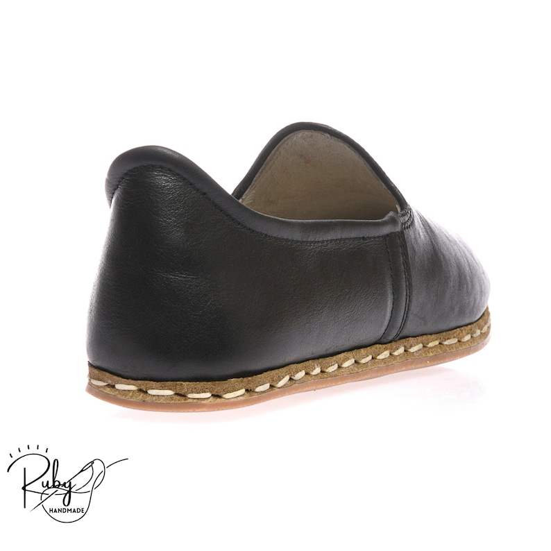 Op de afbeelding: Zwarte leren instappers met een witte gestikte rand rond de zool. De schoenen hebben een afgeronde neus en een platte zool.