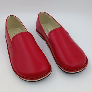 Pode incluir: Um par de sapatos vermelhos de couro com um detalhe de costura branca ao redor da sola. Os sapatos têm um design simples e um visual confortável.