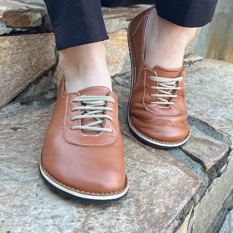 May include: A pair of brown leather lace-up shoes with white laces. The shoes have a white sole and are worn by a person standing on a stone surface.
