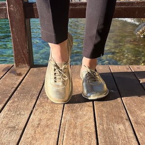 Può includere: Un paio di scarpe in pelle dorate con lacci e punta arrotondata. Le scarpe sono indossate da una persona in piedi su un ponte di legno.