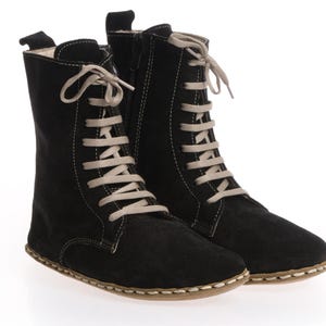 May include: A pair of black suede mid-calf boots featuring light beige laces, a side zip closure, and visible light-coloured stitching along the sole. The boots have a pull tab at the back and a rounded toe design with a flat, flexible sole construction.