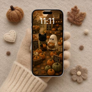 May include: A smartphone displays a pumpkin patch scene with the time 11:11. The phone is held by a person wearing a beige sweater. Surrounding the phone are felt pumpkins, a heart, a leaf, and other decorative elements.