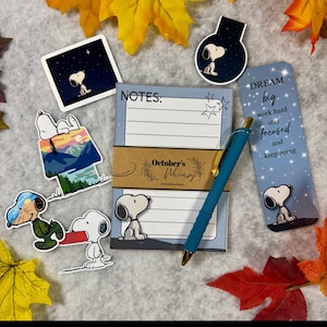 May include: A stationery set featuring a lined notepad with the text NOTES, a blue pen, and several Snoopy stickers. One sticker shows Snoopy on a doghouse, another shows Snoopy with a red book, and a bookmark displays the text DREAM big work hard stay focused and keep going. The set is branded with an October's Whimsy paper band.