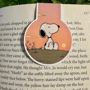 May include: A magnetic bookmark featuring a circular illustration of the cartoon character Snoopy sitting on a grassy hill at sunset. The background shows a soft orange sky with a small sun and simple flowers, while the character is white with black ears.