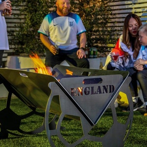 May include: A black metal fire pit shaped like a football with the word "ENGLAND" cut out of the metal. The fire pit is on a green lawn with people in the background wearing football shirts.