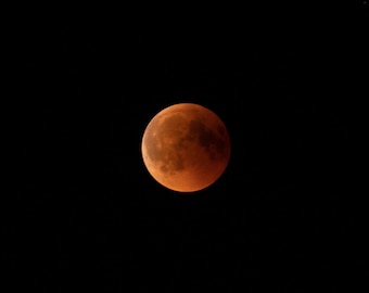 May include: A full, reddish-orange moon against a black background. The moon's surface shows detailed textures and shadows, creating a dramatic celestial image. The image captures the moon in a night sky.