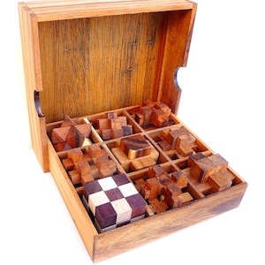 Puede incluir: Una caja de madera con tapa abatible, abierta para revelar nueve rompecabezas de madera diferentes. Los rompecabezas están hechos de varios tipos de madera y están dispuestos en un patrón de cuadrícula.