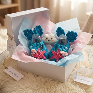 Peut inclure: Une bo&icirc;te cadeau blanche contient une paire de chaussons de b&eacute;b&eacute; bleus au crochet avec des accents d'&eacute;toiles de mer et de coquillages. Les chaussures ont des ailes bleues au crochet. Un jouet en peluche en forme de nuage souriant est pos&eacute; entre les chaussures.