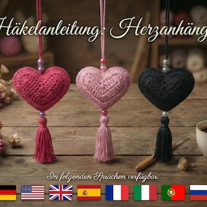 以下が含まれることがあります： ピンクと黒の3つのかぎ針編みのハートの飾りで、それぞれにタッセルが付いており、紐で吊り下げられています。画像にはドイツ語のテキスト、毛糸玉、コーヒーカップが含まれています。さまざまな国の国旗が下にあります。
