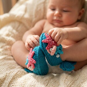 Peut inclure: Une paire de chaussons de b&eacute;b&eacute; en crochet turquoise avec des &eacute;toiles de mer roses et des perles. Les chaussons, en forme de queues de sir&egrave;ne, sont tenus par un b&eacute;b&eacute;. Le b&eacute;b&eacute; est assis sur une couverture en tricot cr&egrave;me.