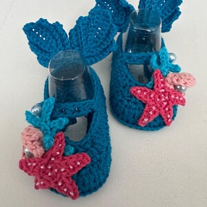 Peut inclure: Une paire de chaussons de b&eacute;b&eacute; au crochet bleu turquoise. Chaque chaussure est orn&eacute;e d'un grand n&oelig;ud, d'une &eacute;toile de mer rose, de coraux bleus et de fausses perles. Ces chaussures faites &agrave; la main ont un th&egrave;me nautique.