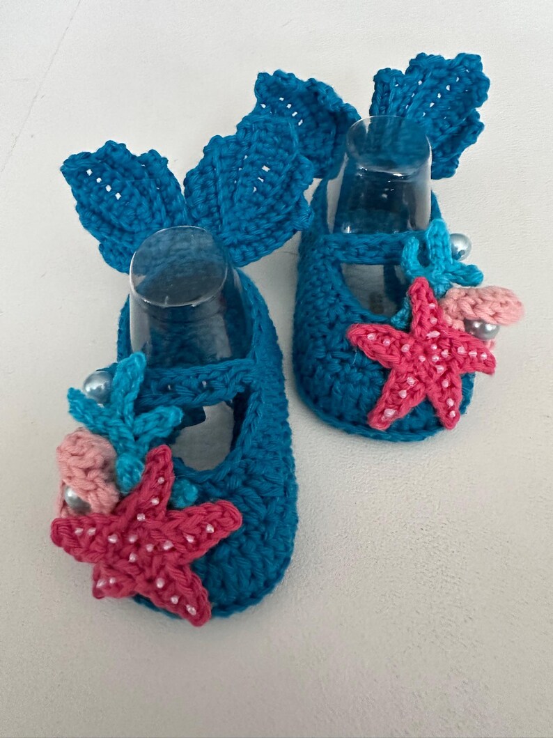Peut inclure: Une paire de chaussons de b&eacute;b&eacute; au crochet bleu turquoise. Chaque chaussure est orn&eacute;e d'un grand n&oelig;ud, d'une &eacute;toile de mer rose, de coraux bleus et de fausses perles. Ces chaussures faites &agrave; la main ont un th&egrave;me nautique.