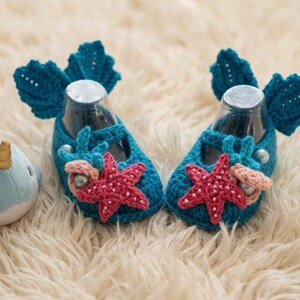 Peut inclure: Une paire de chaussons de b&eacute;b&eacute; en crochet bleu turquoise avec des &eacute;toiles de mer et des coquillages, &agrave; c&ocirc;t&eacute; d'une peluche narval. Les chaussures pr&eacute;sentent un design en forme d'aileron &agrave; l'arri&egrave;re et sont d&eacute;cor&eacute;es d'&eacute;toiles de mer rouges et de petites perles. Le narval est bleu clair avec une corne dor&eacute;e.