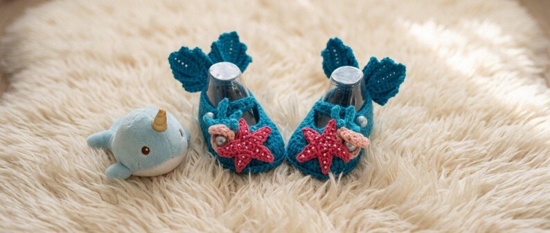 Peut inclure: Une paire de chaussons de b&eacute;b&eacute; en crochet bleu turquoise avec des &eacute;toiles de mer et des coquillages, &agrave; c&ocirc;t&eacute; d'une peluche narval. Les chaussures pr&eacute;sentent un design en forme d'aileron &agrave; l'arri&egrave;re et sont d&eacute;cor&eacute;es d'&eacute;toiles de mer rouges et de petites perles. Le narval est bleu clair avec une corne dor&eacute;e.