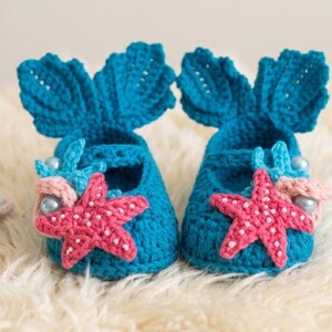 Peut inclure: Une paire de chaussons de b&eacute;b&eacute; turquoise au crochet, avec des accents de queue de sir&egrave;ne et des &eacute;toiles de mer roses. Une petite tortue au crochet aux couleurs pastel est &agrave; c&ocirc;t&eacute; des chaussures, le tout sur une surface blanche et moelleuse. Th&egrave;me nautique.
