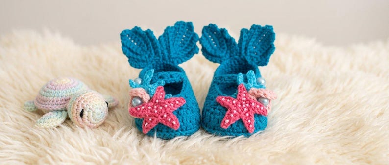 Peut inclure: Une paire de chaussons de b&eacute;b&eacute; turquoise au crochet, avec des accents de queue de sir&egrave;ne et des &eacute;toiles de mer roses. Une petite tortue au crochet aux couleurs pastel est &agrave; c&ocirc;t&eacute; des chaussures, le tout sur une surface blanche et moelleuse. Th&egrave;me nautique.