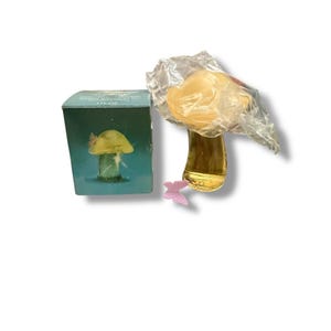 Op de afbeelding: Een decoratieve paddenstoelvormige parfumfles met een gele dop en een heldere, amberkleurige basis. De fles is gedeeltelijk verpakt in plastic. Een blauwgroene doos met een paddenstoelillustratie en een roze vlinder staat naast de fles.