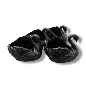 May include: A set of four black swan-shaped decorative bowls. Each bowl features a detailed swan design with a curved neck and a scalloped edge. The bowls are made of a glossy, dark material.