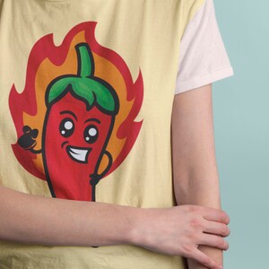 May include: A yellow t-shirt with a cartoon red chili pepper wearing a green hat and surrounded by orange flames. The chili pepper has a smiling face with big eyes.