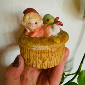 May include: A ceramic trinket box with a whimsical design featuring a small elf laying on a tree stump. The elf is wearing a red and white hat and a red and white dress. The tree stump is brown and has a textured surface.
