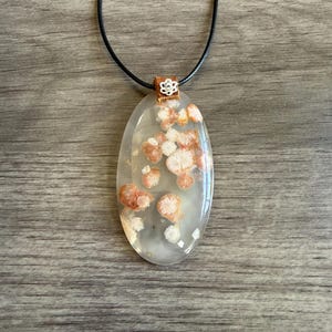 May include: An oval-shaped pendant featuring a translucent, milky white stone with embedded clusters of reddish-orange, flower-like inclusions. The pendant is suspended from a thin black cord by a copper-colored bail with a small silver flower accent.