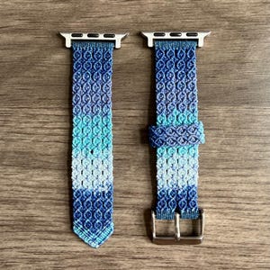 May include: A two-piece watch band featuring a macrame knot pattern in a gradient of blue, teal, and white. The band includes silver-toned metal connectors at the top and a silver-toned metal buckle on the lower section for secure attachment.