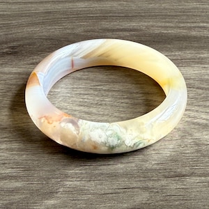 May include: A round, polished gemstone bracelet with a mix of colors, including white, yellow, orange, and green. The bracelet is smooth and has a natural, organic appearance. The bracelet is a piece of jewelry.