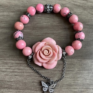 May include: A pink gemstone bracelet with a rose charm and a butterfly pendant. The bracelet is made of pink and white stones and has a black metal clasp.