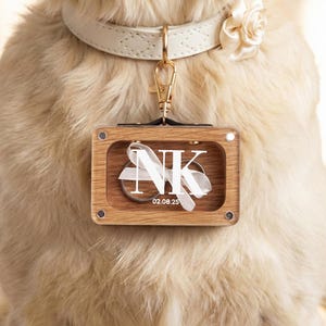 Könnte beinhalten: Ein personalisiertes Haustier-Tag mit Holzrahmen und klarer Acrylfront. Das Tag zeigt die Initialen "NK" mit einem weißen Schleifendesign und dem Datum "02.08.25". Es ist an einem cremefarbenen Lederhalsband mit einem goldenen Verschluss und einer Stoffrose befestigt.