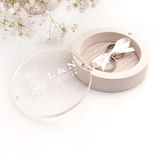 Könnte beinhalten: Eine transparente Acryl-Ringbox mit einem gravierten Blumenmuster und dem Text "L&N 20.07.24". Die Box ist geöffnet und eine weiße Ringbox mit zwei goldenen Ringen und einem weißen Band ist im Inneren sichtbar.