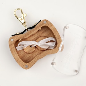 May include: A wooden ring box with a gold-tone metal lobster clasp attached to a black fabric strap. The box has a light wood grain finish, a hollow center containing two wooden rings tied with a white ribbon, and a clear acrylic lid.