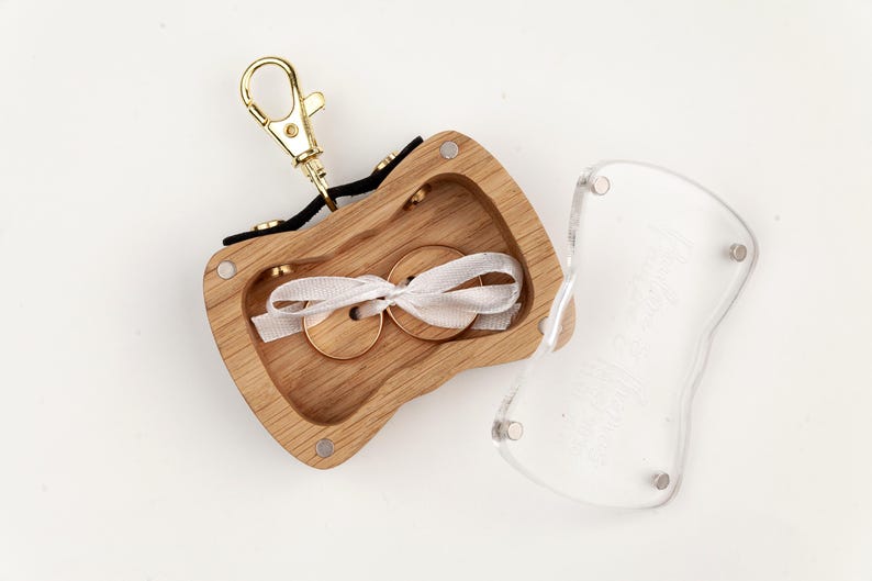 May include: A wooden ring box with a gold-tone metal lobster clasp attached to a black fabric strap. The box has a light wood grain finish, a hollow center containing two wooden rings tied with a white ribbon, and a clear acrylic lid.