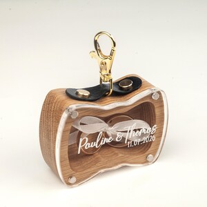 May include: A wooden ring box with a clear acrylic front panel featuring the white printed text Pauline & Thomas 11.07.2026. Inside, two gold rings are tied with a white ribbon. The box has a black leather strap with gold snaps and a gold clasp.
