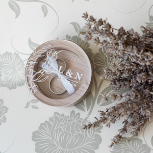 May include: A wooden ring box with a clear lid, featuring a floral design and the initials "L&N" with the date "2024". Two silver rings are inside the box, with a white ribbon.