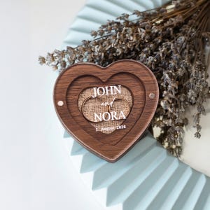 A wooden heart-shaped box with the names 'JOHN and NORA' engraved on it, placed on a white surface.