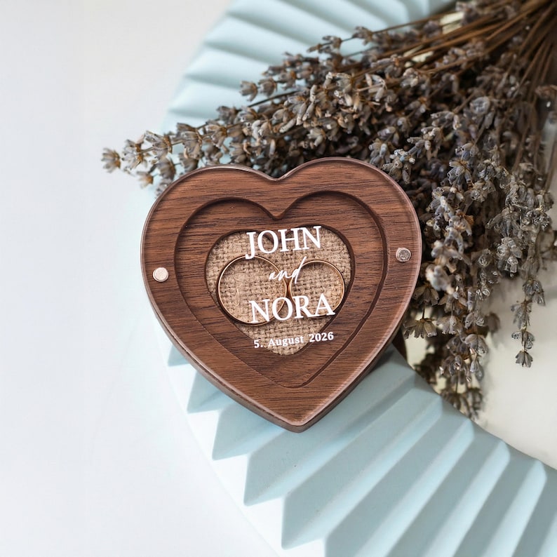 A wooden heart-shaped box with the names 'JOHN and NORA' engraved on it, placed on a white surface.