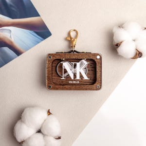 May include: A wooden keychain with a gold clasp, featuring a clear window displaying two rings and the initials "NK" with the date "02.08.25". The keychain has a rectangular shape with rounded corners.