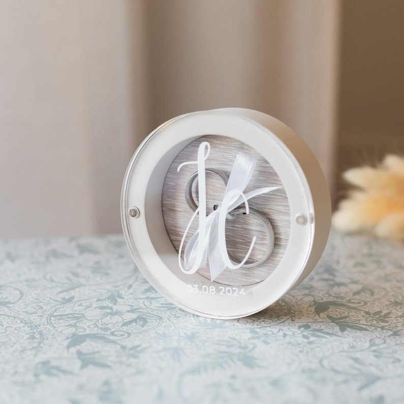 May include: White round ring box with a clear lid. The box has a wood-like interior with a white ribbon and the initials "J & C" in a cursive font. The date "03.08.2024" is printed on the bottom of the box.