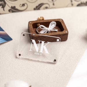 May include: A clear acrylic sign with the initials "NK" and the date "02.08.25" in white lettering. A small, dark wooden ring box with a white ribbon and a ring inside is in the background. The items are on a textured, off-white surface.