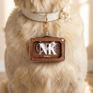 Op de afbeelding: Een hondenhalsband met een crèmekleurige leren riem en een crèmekleurige roos. Een houten label met de initialen "NK" en de datum "02.08.25" is met een goudkleurige sluiting aan de halsband bevestigd.