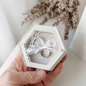 May include: White hexagonal ring box with a clear lid. The box has a floral design and the text "V&P 12.07.2024" engraved on the lid. Two silver rings are inside the box.