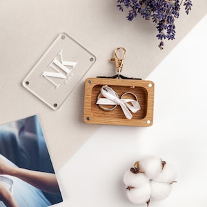 Puede incluir: Un joyero de madera con cierre dorado, que contiene dos anillos de plata atados con una cinta blanca. Una etiqueta de acrílico transparente con las iniciales "NK" y la fecha "02.08.23" está al lado de la caja.